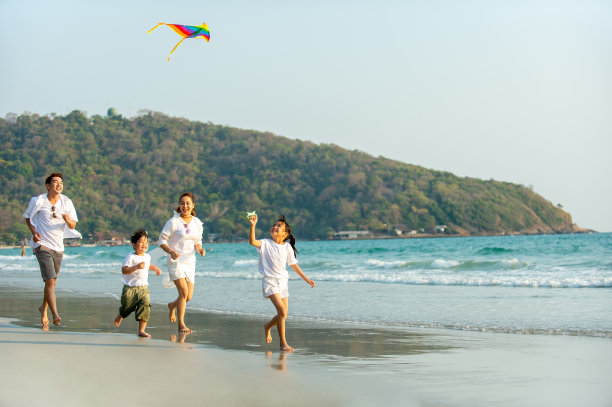 快乐的亚洲家庭父母和两个孩子跑步和玩风筝一起在夏天的时间在海滩上日落。父亲，母亲和孩子们放松和享受夏季户外生活方式假期。图片下载