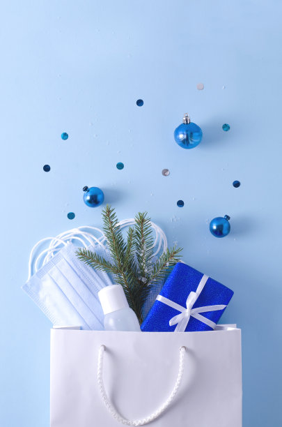 圣诞概念，新年礼物在冠状病毒。白色袋子与保护性一次性口罩，礼物，防腐剂，圣诞树和圣诞玩具在一个蓝色垂直背景与平lay图片下载
