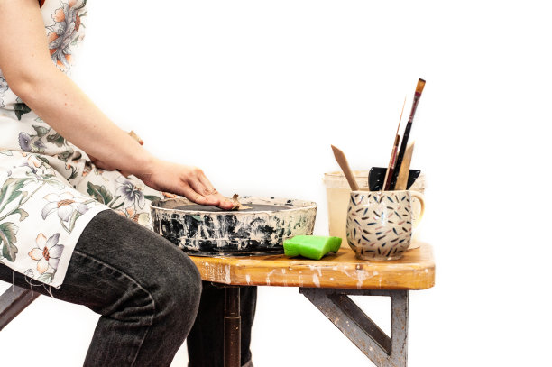 女孩穿着黑色牛仔裤，红色衬衫和白色围裙坐在木凳上做陶器，工具就在附近，白色孤立的形象图片下载