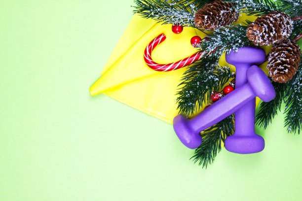 圣诞健身。健康和积极的生活方式贺卡的概念。紫色的哑铃，黄色的橡皮筋，糖果和粉红色的冷杉树图片下载
