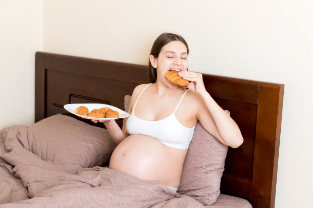 一名孕妇躺在床上吃着家里盘子里的牛角面包。不健康的饮食。怀孕期间控制体重图片下载