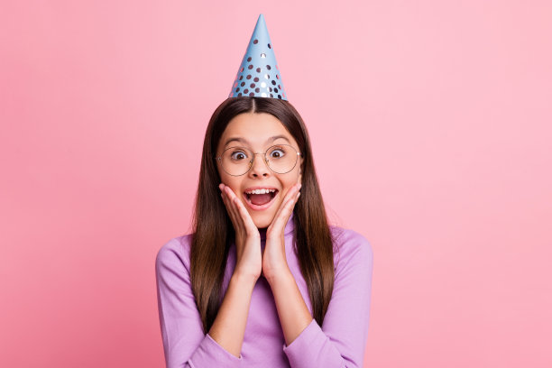 照片惊讶的小女孩在生日帽触摸双手脸颊穿紫色套头衫孤立在柔和的颜色背景图片下载