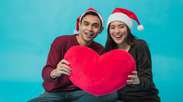 年轻的亚洲情侣戴着圣诞老人的帽子。手握红色心形枕头孤立在蓝色背景在工作室拍摄。圣诞节庆祝情人的概念。图片下载