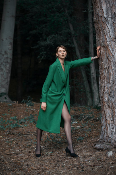 时尚的年轻女子在绿色大衣摆姿势在森林附近的树图片下载