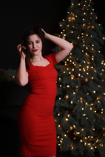 性感迷人的黑发在红色紧身连衣裙附近的圣诞树。黑暗的照片。工作室拍摄。情感上的女演员。图片下载