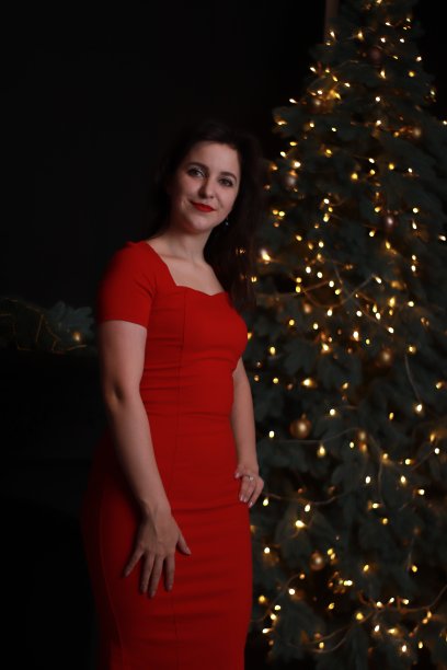 性感迷人的黑发在红色紧身连衣裙附近的圣诞树。黑暗的照片。工作室拍摄。情感上的女演员。图片下载