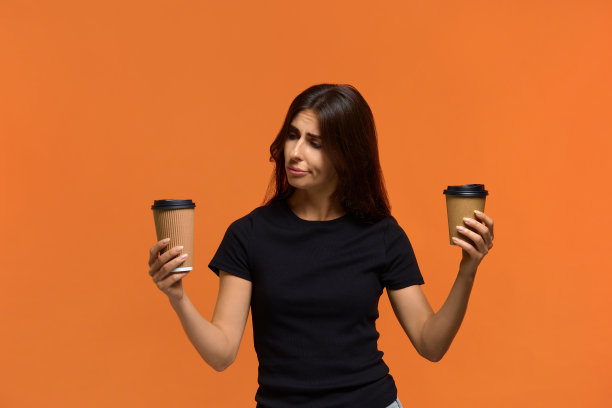该选择什么。一名身穿黑色t恤的白人女性在两种一次性杯子里选择饮料。孤立在橙色背景上。图片下载