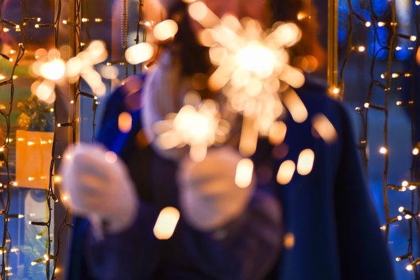 在新年模糊的灯光背景下，一个穿着蓝色外套、戴着手套的笑着的女孩手里燃烧着烟花。圣诞和新年的背景。图片下载