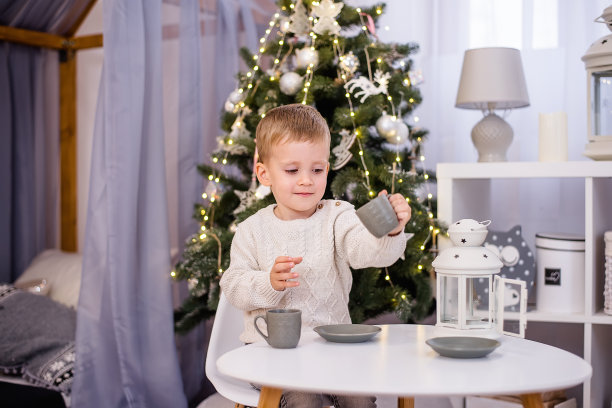 在孩子们的房间里，一个穿着白色套头衫的小男孩在他的白色桌子旁喝茶，用灰色的杯子喝可可。背景是一棵挂着彩灯花环的圣诞树。本空间图片下载