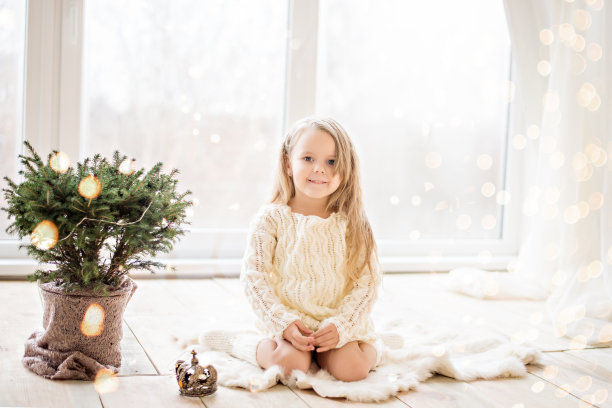 小女孩穿着白色针织毛衣连衣裙，在全景窗口边玩得很开心，跳舞，用彩灯花环装饰一个花盆里的圣诞树。冬天休息，等待新的一年图片下载