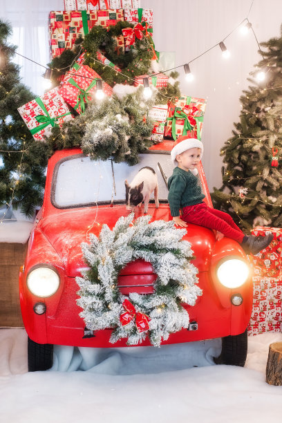 一个小男孩戴着圣诞老人的帽子，坐在一辆红色汽车的引擎盖上，车上有一只迷你猪，圣诞树上装饰着礼物盒，雪松花环，彩灯花环。拷贝空间，新的一年图片下载