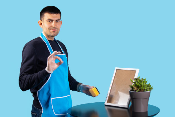 一位穿着围裙、戴着手套的英俊男子，擦去他的热情，心满意足地看着镜头。照片在蓝色的ok标志上。性别刻板印象的概念图片下载