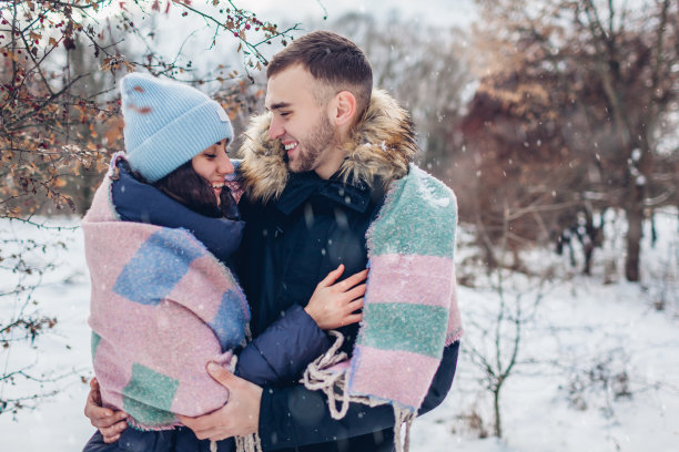 年轻的情侣在冬天的森林里散步和欢笑。人们盖着毯子在飘落的雪下玩图片下载
