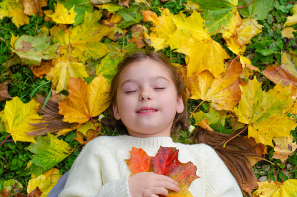 一个穿着白色针织连衣裙的小女孩微笑着躺在落叶上。秋天公园里明亮的黄色、橙色的枫叶。图片下载