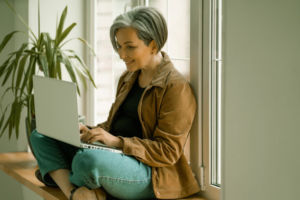 成熟女性会面带微笑地看着网络摄像头，参加虚拟视频聊天、用电脑工作或背靠窗户坐着。侧视图。右边的复制空间图片下载
