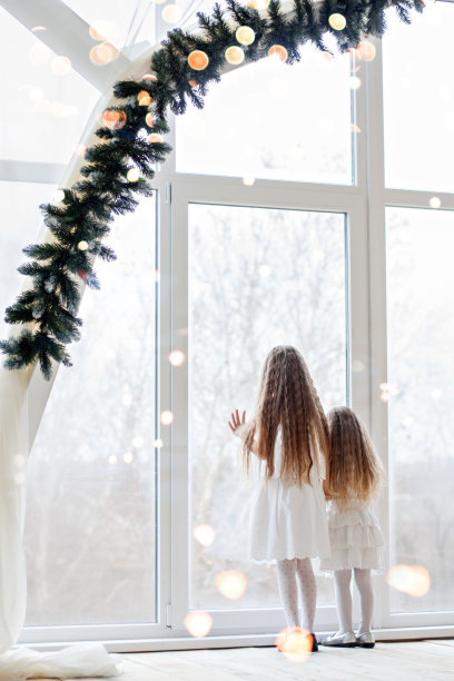 两个金发碧眼的姐妹，穿着白色节日礼服，看着挂满圣诞树树枝的全景窗口，期待着新年的奇迹和圣诞老人。女孩牵手。图片下载