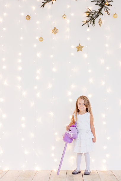 一个金发小女孩穿着白色的节日服装，背景是黄色的散焦从圣诞树枝花环灯。新年贺卡。婴儿开心地笑了，手里拿着一根紫色的玩具马杖。副本的空间。图片下载