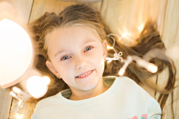 快乐的女孩躺在木地板上，头发上戴着一个明亮的圣诞彩灯花环。微笑着，看着镜头。从上面拍摄，特写肖像。拷贝空间，贺年卡图片下载