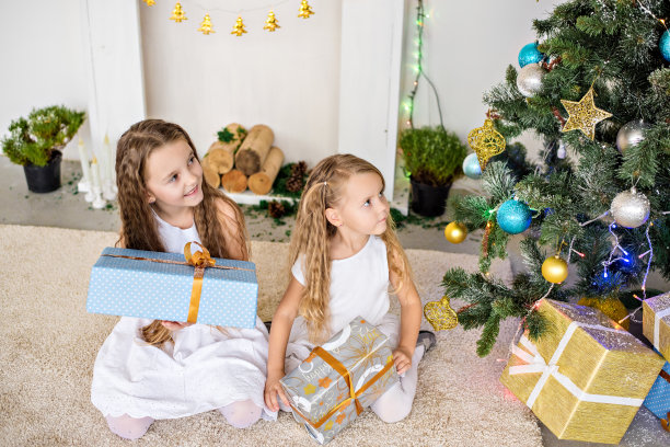 两个穿着白裙子的金发小女孩在壁炉和圣诞树旁玩盒子。姐妹们有乐趣，拥抱，大笑，打开圣诞老人的礼物。新年贺卡。副本的空间。友谊图片下载