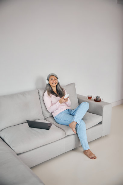 美丽微笑的女人坐在客厅的灰色沙发上，手里拿着手机图片下载