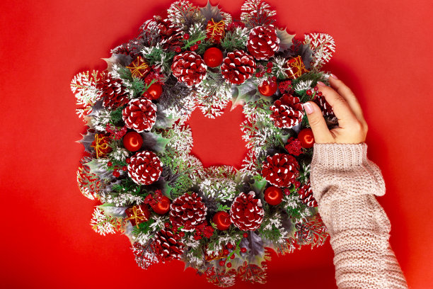 一位穿着针织毛衣的女性手在红色背景上的圆锥形、浆果和气球组成的圣诞花环上添加了冷杉圆锥形图片下载