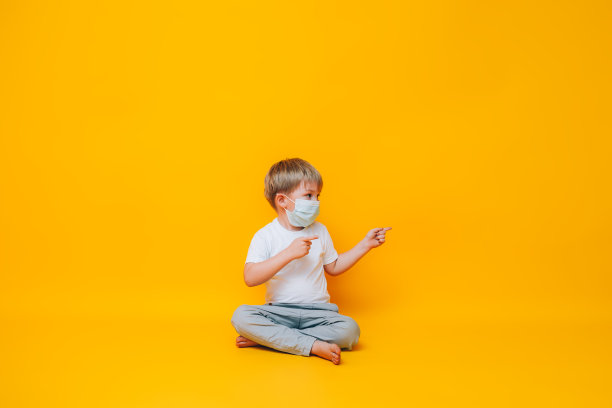 可爱的男孩在黄色背景点用他的手向一边，照片为广告产品医疗口罩在脸上，流行图片下载