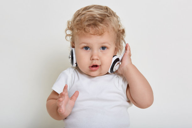 婴儿戴着耳机直视镜头，手一直拿着耳机，面部表情好奇，孩子穿着t恤，在白色背景下摆出孤立的姿势。图片下载