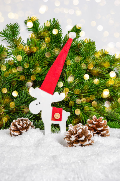 白色圣诞驯鹿在红色的圣诞帽和松果在雪与冷杉树和发光的散焦灯作为背景。节日贺卡。圣诞快乐，恭贺新禧图片下载