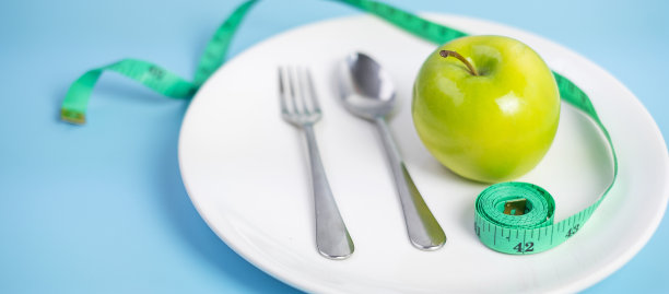 俯视图勺子和叉子，绿色的苹果在白色的陶瓷盘子和绿色的卷尺在蓝色的背景。节食、减肥、肥胖和食物控制的概念图片下载