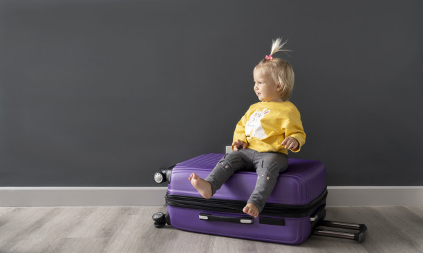 疫情后，坐在紫色行李箱上等待旅行的可爱小孩子，灰色背景，留白图片下载