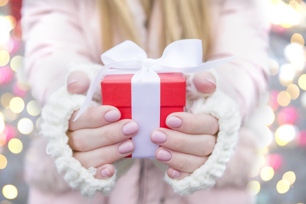 女人手拿圣诞红礼盒与弓在假日背景与灯光散景，特写。圣诞和新年的问候季节概念图片下载