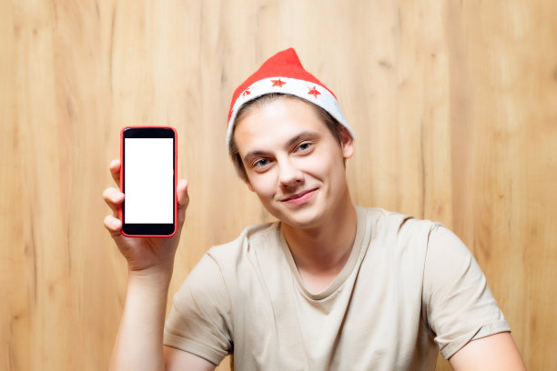 现代人在室内戴着红色的圣诞老人帽。微笑着用手机的人的肖像。男孩正在显示空白屏幕的设备复制空间模板和模拟图片下载