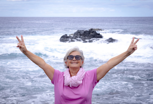 前面的观点，一个白发的资深妇女穿着粉红色的享受自由与她的手臂伸出在海边。寒假里积极退休，海阔天空图片下载