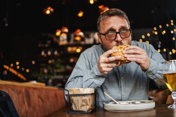 蓄着胡须的中年饥饿男子坐在餐馆里吃着美味的汉堡。图片下载