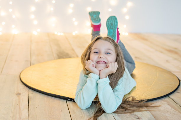 一个漂亮的金发小女孩躺在木地板上一个金色的圆圈上，背景是一堆模糊的黄色圣诞花环。新年贺卡。蹒跚学步的运动。复制空间，喜庆纹理图片下载
