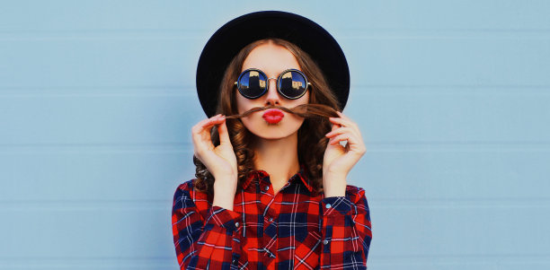 肖像近距离的有趣的女人显示胡子，她的头发吹红色的嘴唇戴着黑色的圆帽子在蓝色的背景图片下载