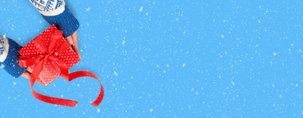 戴着手套的手拿着一个红色的礼品盒，上面有红色的丝带，背景是蓝色的雪。图片下载