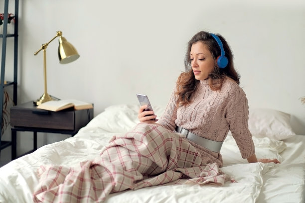 年轻快活的女人躺在床上，用手机拖着脚唱着歌。放松的女孩在家里用耳机听音乐。快乐的女人在不经意间享受着卧室里的音乐，同时发着手机短信图片下载