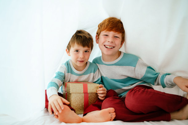 小兄弟姐妹们在白色的床单下打开圣诞礼物盒的水平视图。图片下载