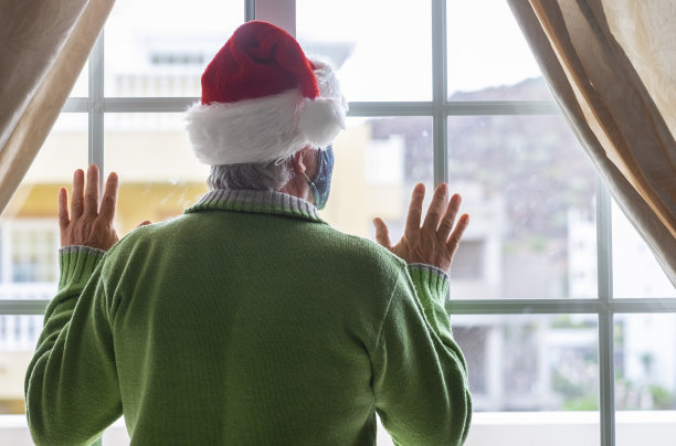 后视图的一个老人戴着圣诞老人帽，绿色毛衣和面罩呆在家里的窗户后面看出去。因covid-19冠状病毒而被封锁的孤独概念图片下载