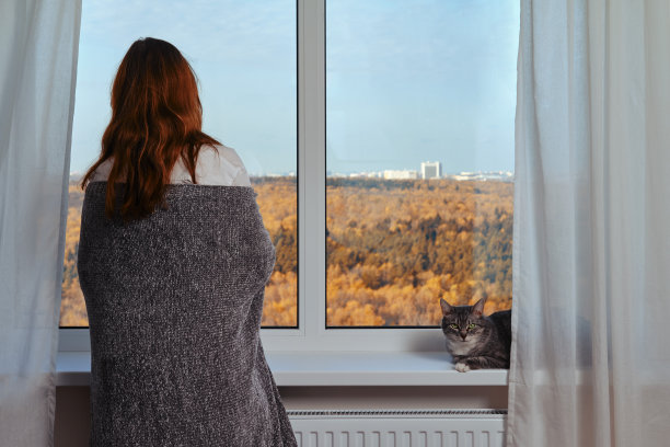 窗边一个成年女子裹着灰色毯子，旁边是一只猫。窗外秋色自然，家舒适图片下载