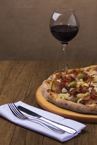 葡萄牙披萨，由火腿、豌豆蛋、棕榈心、意大利辣香肠、洋葱、马苏里拉奶酪和培根制成。一杯红酒。一张白色餐巾纸上竖着刀叉的美食照片。图片下载