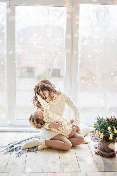 圣诞树旁，美丽的妈妈和快乐的女儿穿着毛衣，在全景窗口玩耍。一个年轻女子微笑着拥抱小女孩。新年贺卡。本空间图片下载