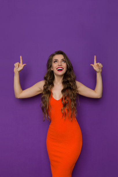 快乐的年轻女人在紧身橙色连衣裙指向头顶和向上看图片下载