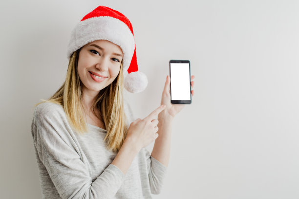 一名年轻女子在白色墙壁前的肖像，她戴着圣诞老人的帽子，拿着手机和显示设备屏幕图片下载