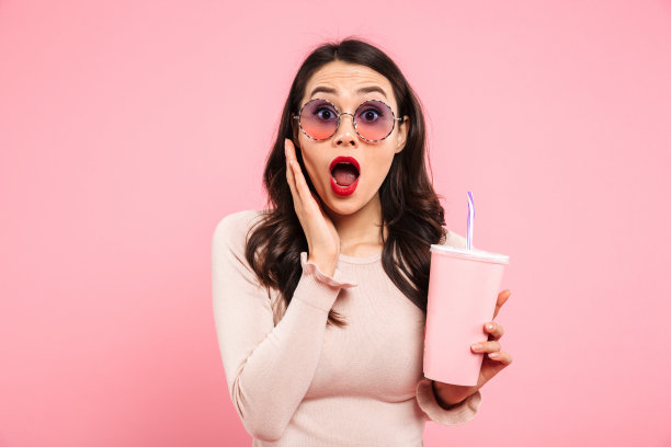 照片激动的成年女孩与长黑发在圆眼镜拿着冷饮在纸杯，孤立在粉红色的背景图片下载