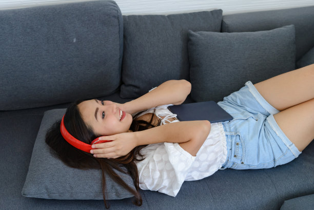 美丽的年轻亚洲女人的肖像戴着耳机听音乐，而放松在家里的沙发上。在家放松。图片下载