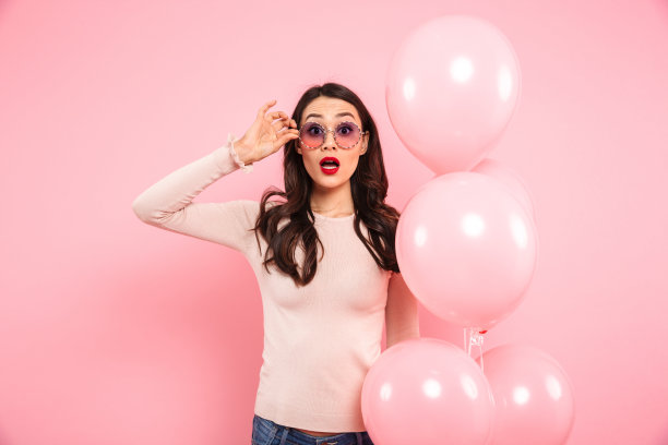 美丽的成年女孩的照片与长黑发在圆圆的眼镜拿着许多气球和表达兴奋，孤立在粉红色的背景图片下载