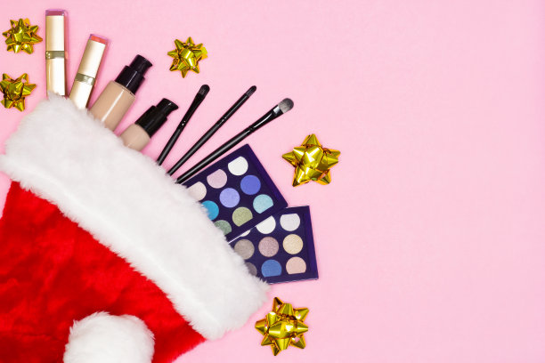 化妆产品设置在圣诞老人的帽子与闪亮的礼物蝴蝶结在粉红色的背景-俯视图。化妆化妆作为圣诞和新年礼物的概念。本空间图片下载