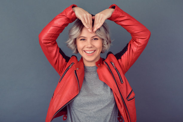 年轻女子身穿皮夹克孤立地站在灰色的墙上，双手呈心形图片下载